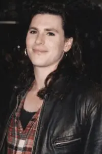 Woman with curly hair wearing a leather jacket and patterned shirt.